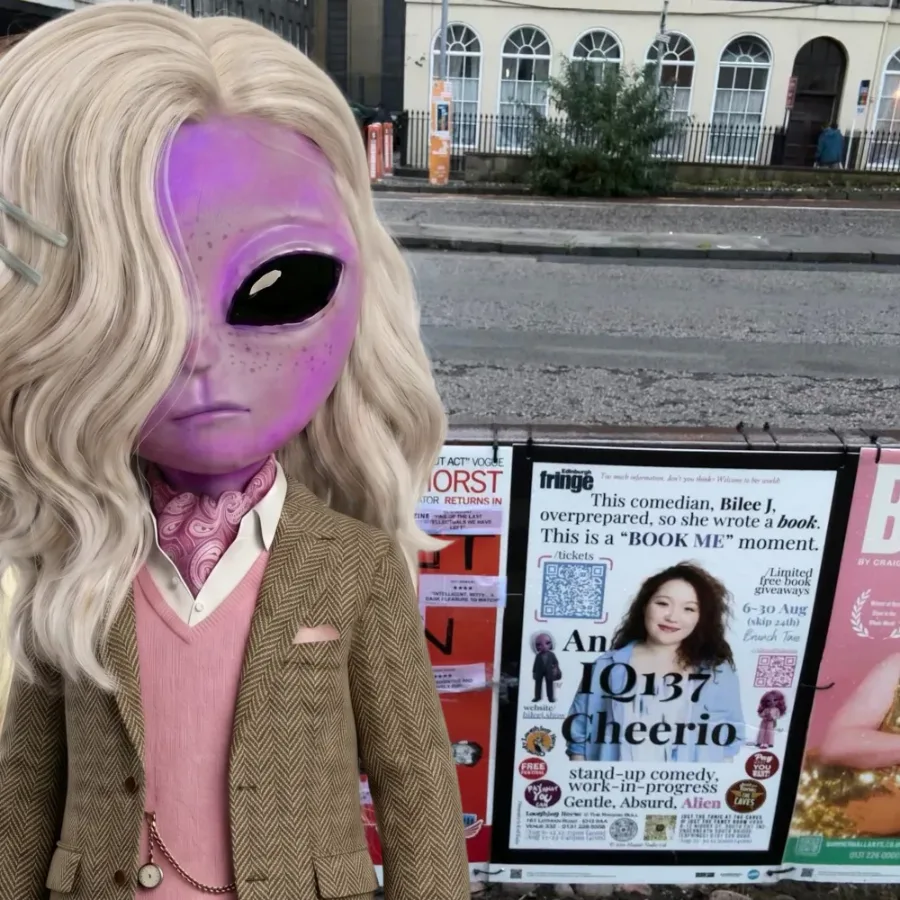 Felix, a lilac alien with long wavy blonde hair, a tweed jacket and a pink cravat, standing beside Bilee J's Fringe poster for An IQ137 Cheerio on street railings