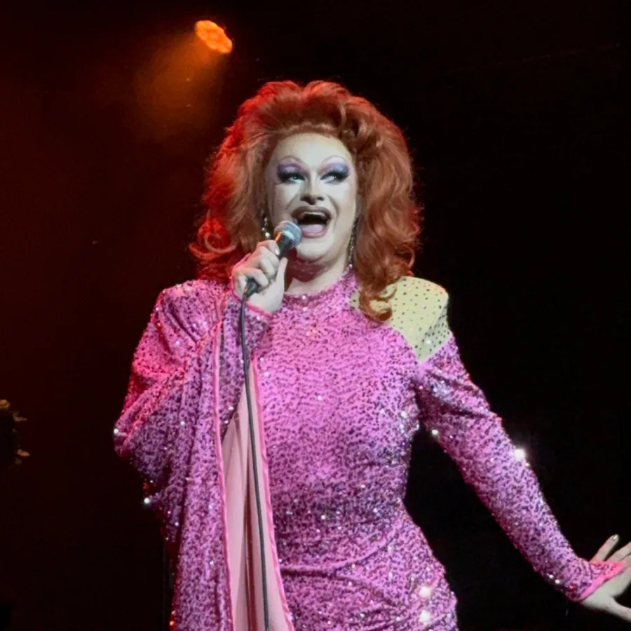 Kate Butch on stage in a pink sequinned gown, performing with a microphone under a spotlight