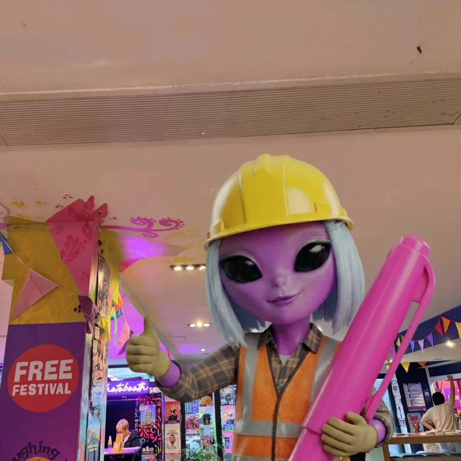 Rebecca the alien in a yellow hard hat, holding a giant pink pen and giving a thumbs up inside a Free Festival venue