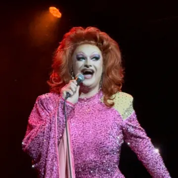 Kate Butch on stage in a pink sequinned gown, performing with a microphone under a spotlight